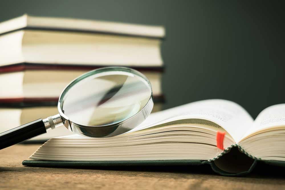 Magnifying glass on the opened book on the table, with pile of books on background.